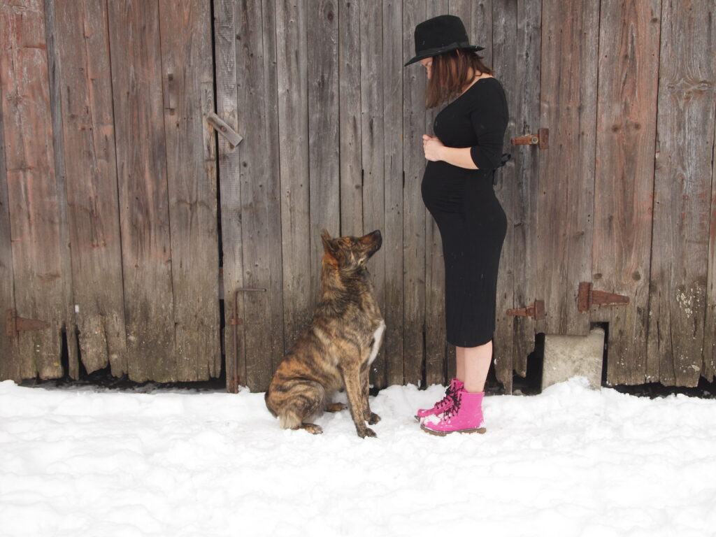 Těhotná Julia v černých šatech a s kloboukem. Před ní sedí pes.
