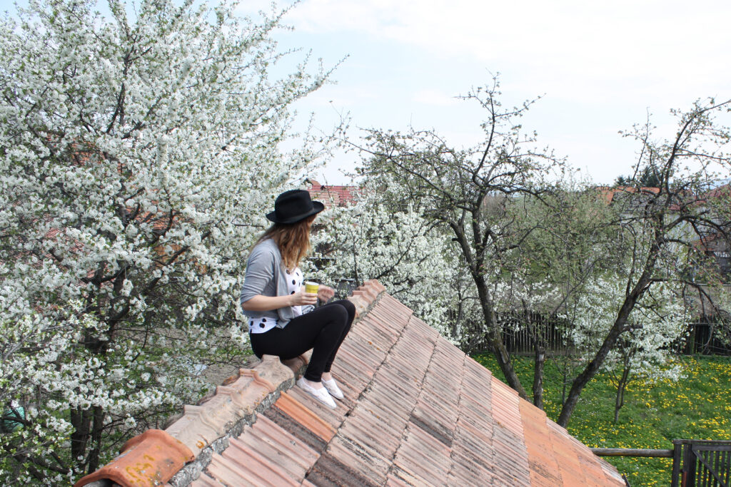 Júlia sedící na střeše s pitím, v klobouku, na pozadí rozkvetlé ovocené stromy.